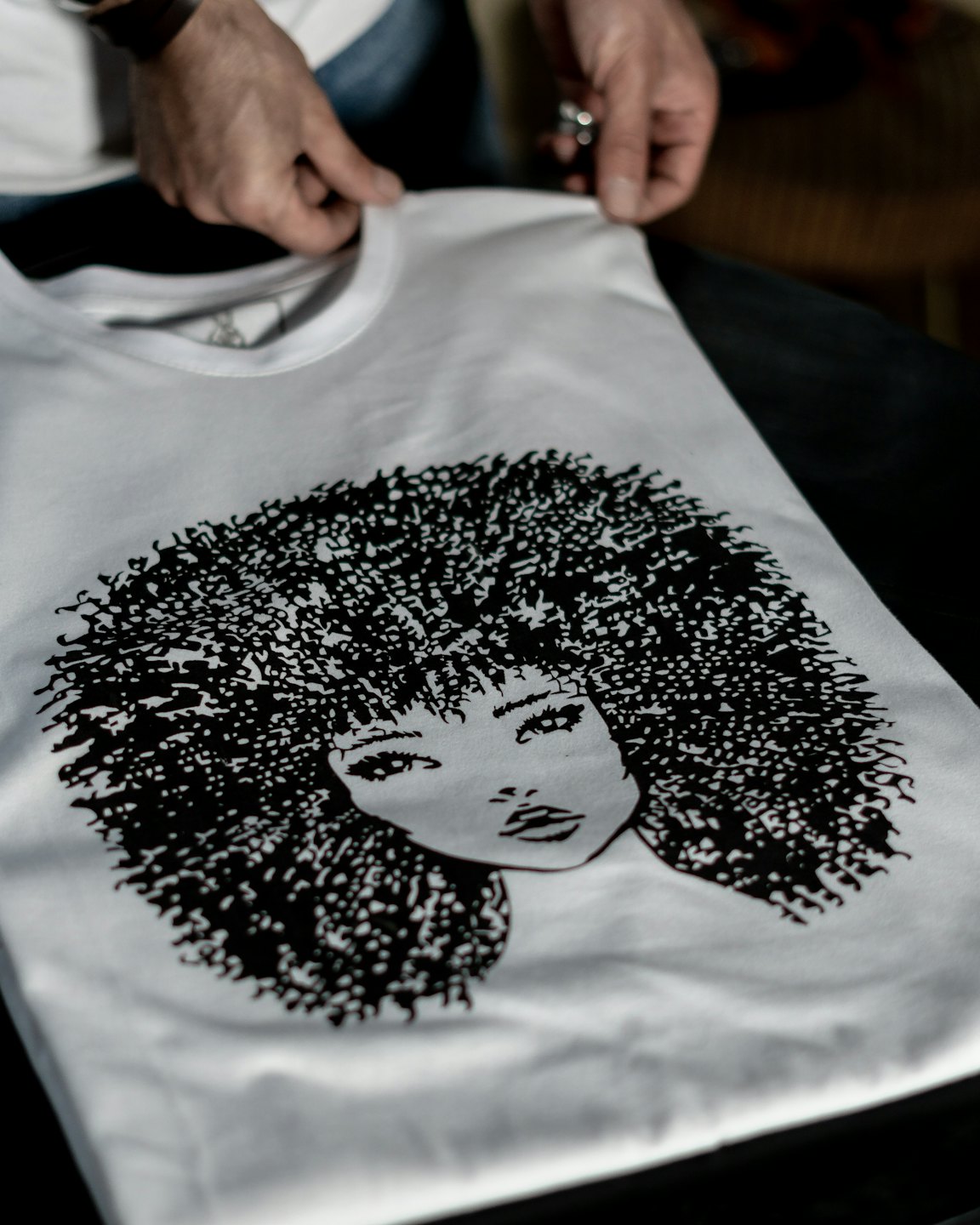 Handen inspecteren een wit T-shirt met een zwarte krulharenillustratie.
